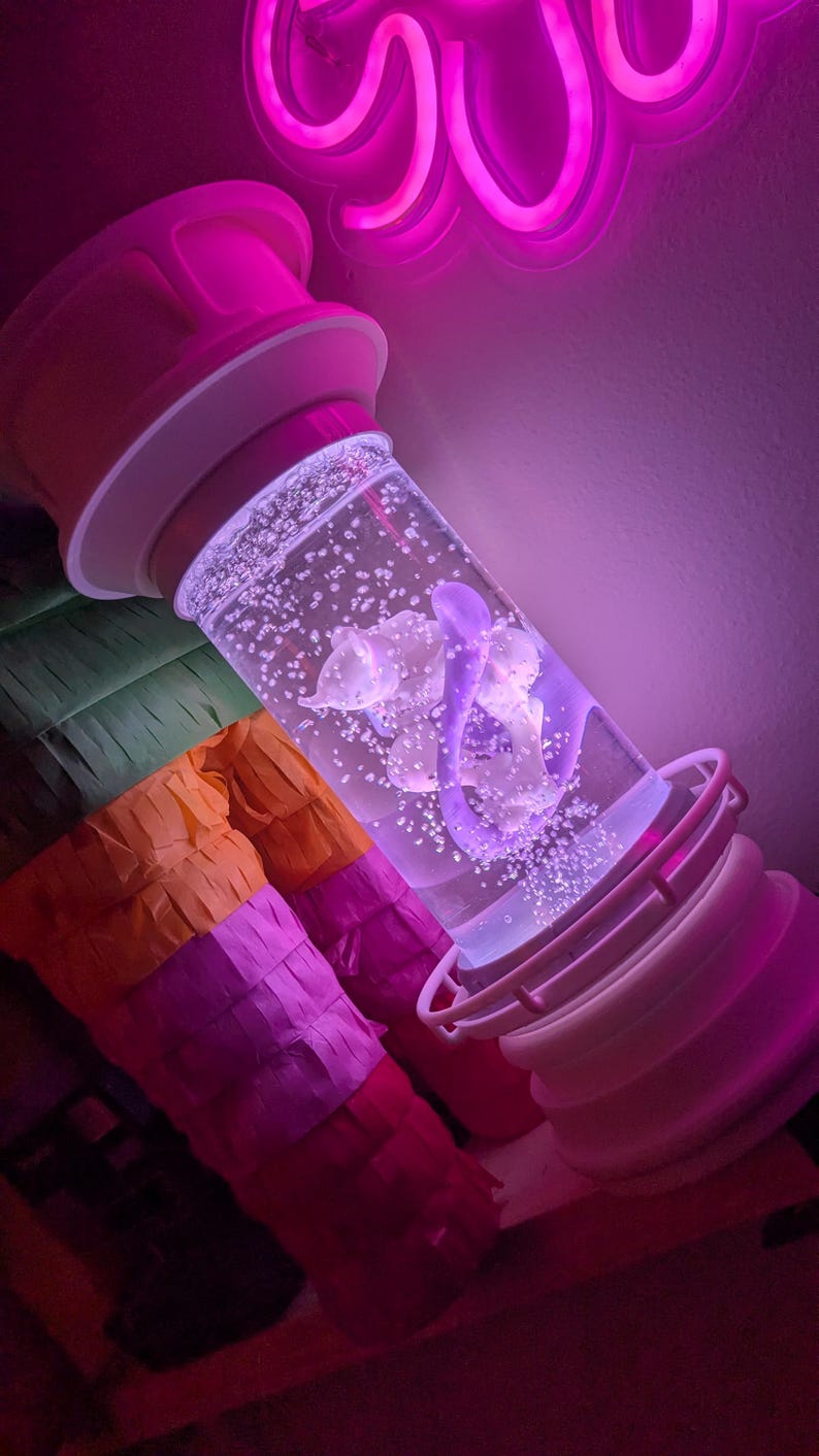 May include: A cylindrical liquid-filled sensory lamp featuring a white base and cap, containing a purple floating shape and numerous small air bubbles. The lamp is illuminated by a vibrant pink neon light, casting a glow on the clear liquid.