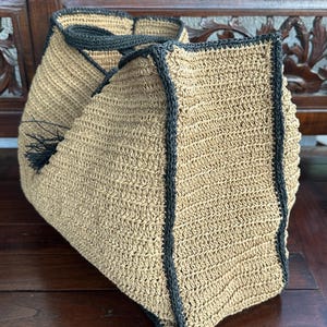 K&ouml;nnte beinhalten: Eine gro&szlig;e, beige geh&auml;kelte Tragetasche mit schwarzem Besatz. Die Tasche hat einen Quasten an der Seite und ist aus Naturfaser gefertigt.