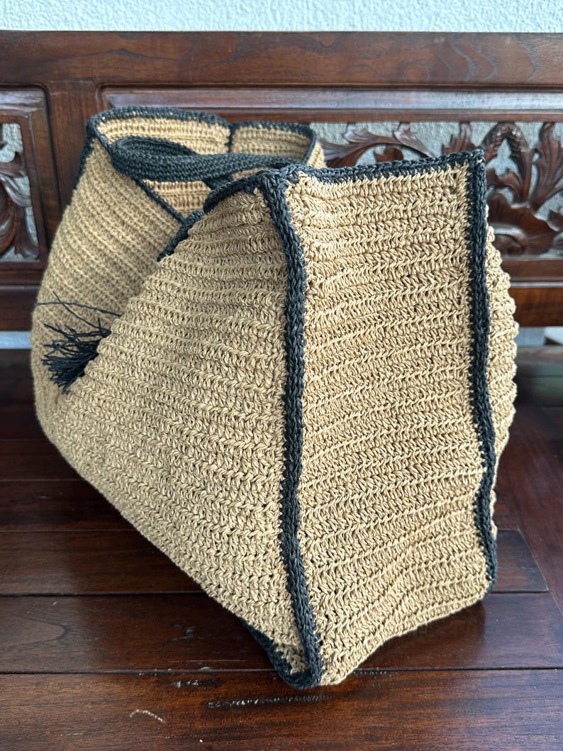 K&ouml;nnte beinhalten: Eine gro&szlig;e, beige geh&auml;kelte Tragetasche mit schwarzem Besatz. Die Tasche hat einen Quasten an der Seite und ist aus Naturfaser gefertigt.