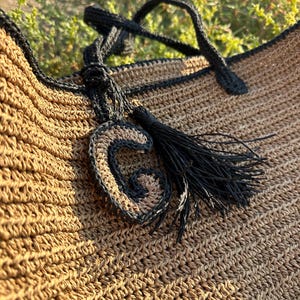 K&ouml;nnte beinhalten: Eine braune geh&auml;kelte Tragetasche mit schwarzem Besatz und einem schwarzen Quasten. Die Tasche hat einen schwarzen geh&auml;kelten Buchstaben "G" daran befestigt.