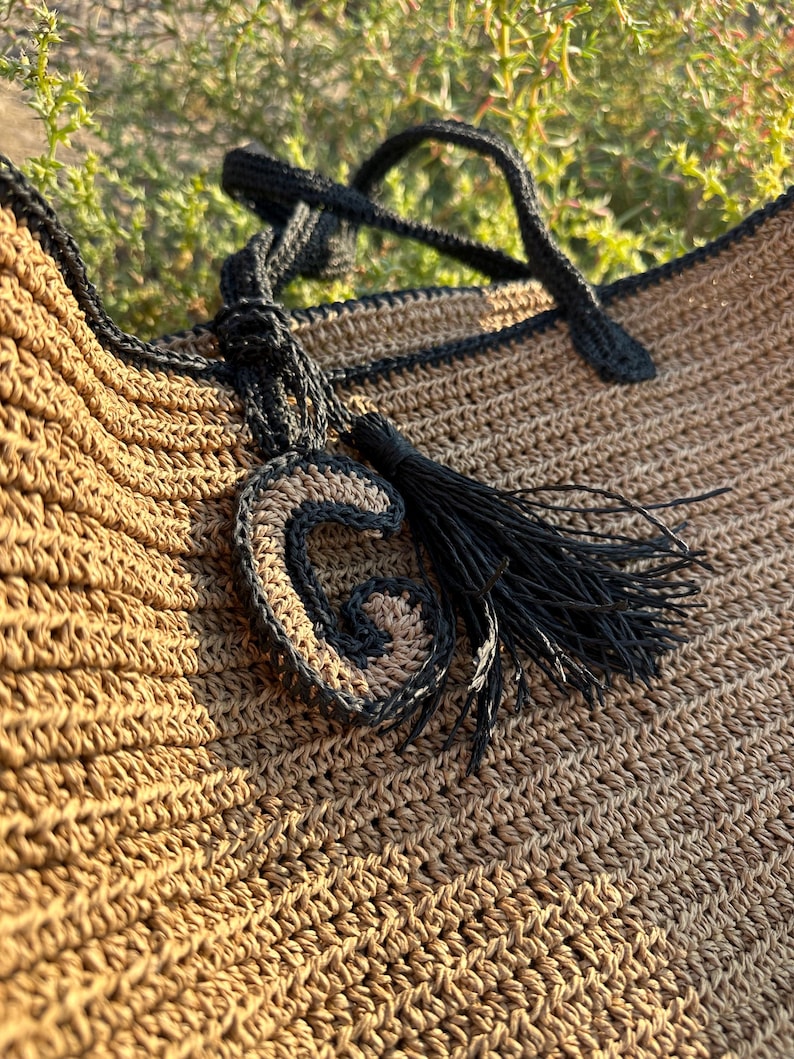 K&ouml;nnte beinhalten: Eine braune geh&auml;kelte Tragetasche mit schwarzem Besatz und einem schwarzen Quasten. Die Tasche hat einen schwarzen geh&auml;kelten Buchstaben "G" daran befestigt.