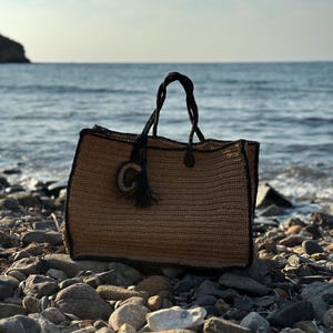 K&ouml;nnte beinhalten: Eine braune und schwarze gewebte Tragetasche mit einem schwarzen Quasten. Die Tasche liegt auf einem steinigen Strand.