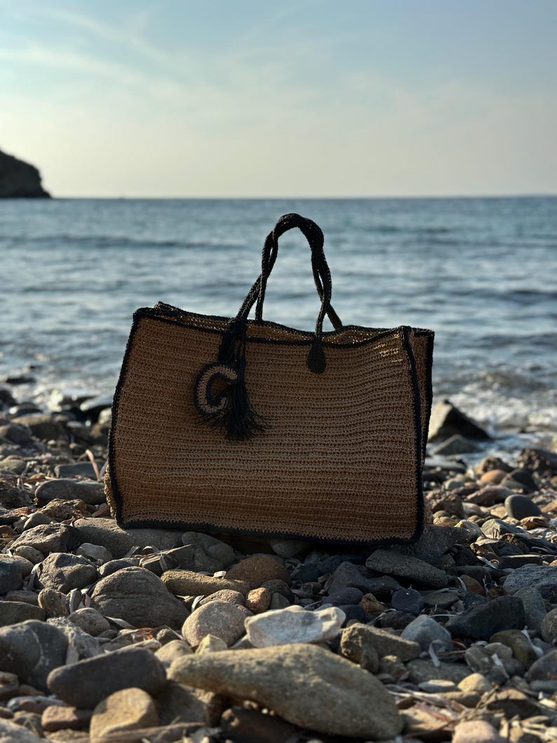K&ouml;nnte beinhalten: Eine braune und schwarze gewebte Tragetasche mit einem schwarzen Quasten. Die Tasche liegt auf einem steinigen Strand.