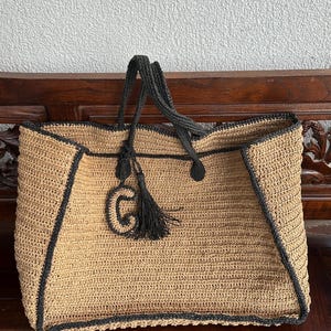 K&ouml;nnte beinhalten: Eine gro&szlig;e, beige geh&auml;kelte Tragetasche mit schwarzem Besatz und einem schwarzen Quasten. Die Tasche hat einen schwarzen gestickten Buchstaben "G" auf der Vorderseite.