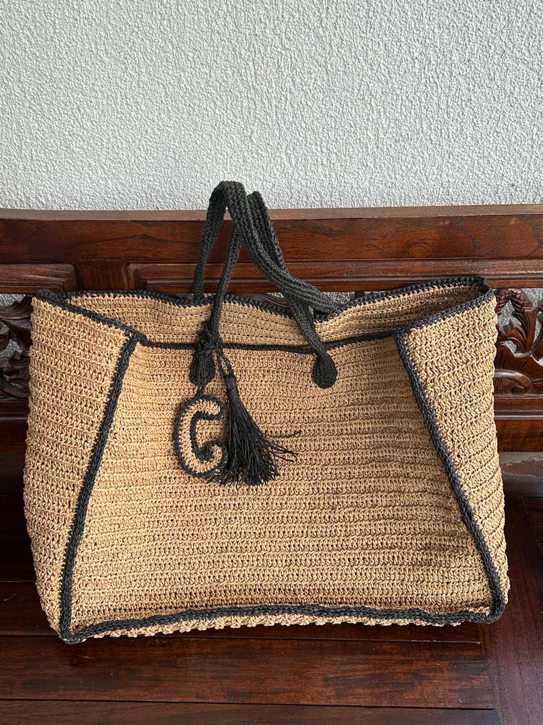 K&ouml;nnte beinhalten: Eine gro&szlig;e, beige geh&auml;kelte Tragetasche mit schwarzem Besatz und einem schwarzen Quasten. Die Tasche hat einen schwarzen gestickten Buchstaben "G" auf der Vorderseite.