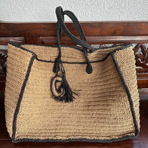 K&ouml;nnte beinhalten: Eine gro&szlig;e, beige geh&auml;kelte Tragetasche mit schwarzem Besatz und Henkeln. Die Tasche hat einen schwarzen Quastenanh&auml;nger an der Vorderseite.