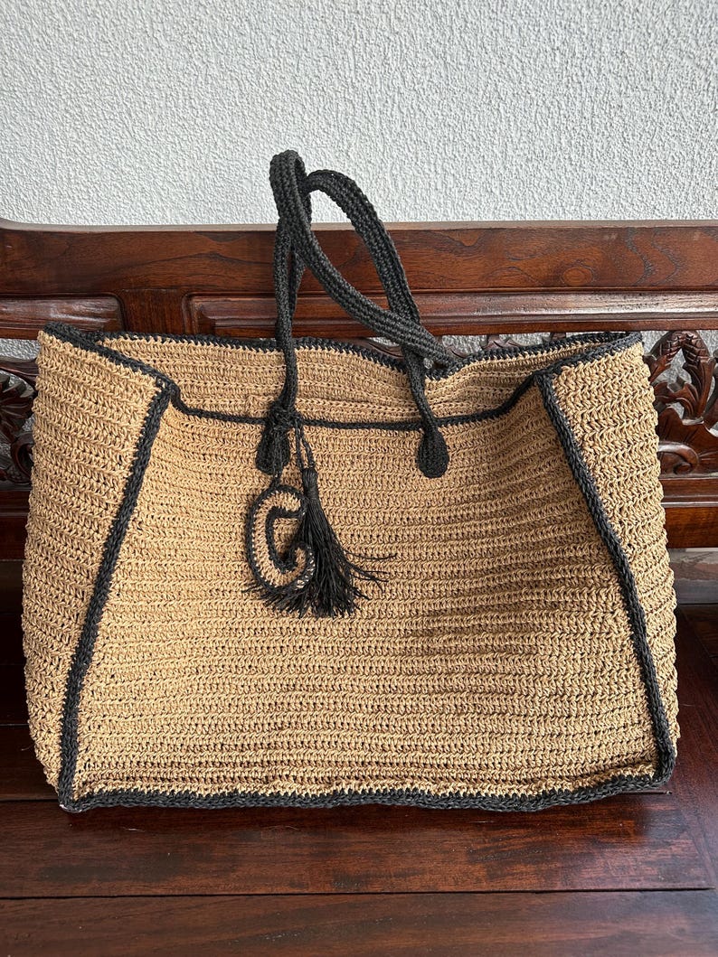 K&ouml;nnte beinhalten: Eine gro&szlig;e, beige geh&auml;kelte Tragetasche mit schwarzem Besatz und Henkeln. Die Tasche hat einen schwarzen Quastenanh&auml;nger an der Vorderseite.