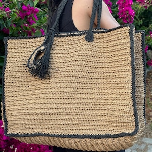 K&ouml;nnte beinhalten: Eine gro&szlig;e, gewebte Tragetasche mit schwarzem Besatz und einem schwarzen Quasten. Die Tasche ist aus einem nat&uuml;rlichen stroh&auml;hnlichen Material gefertigt.