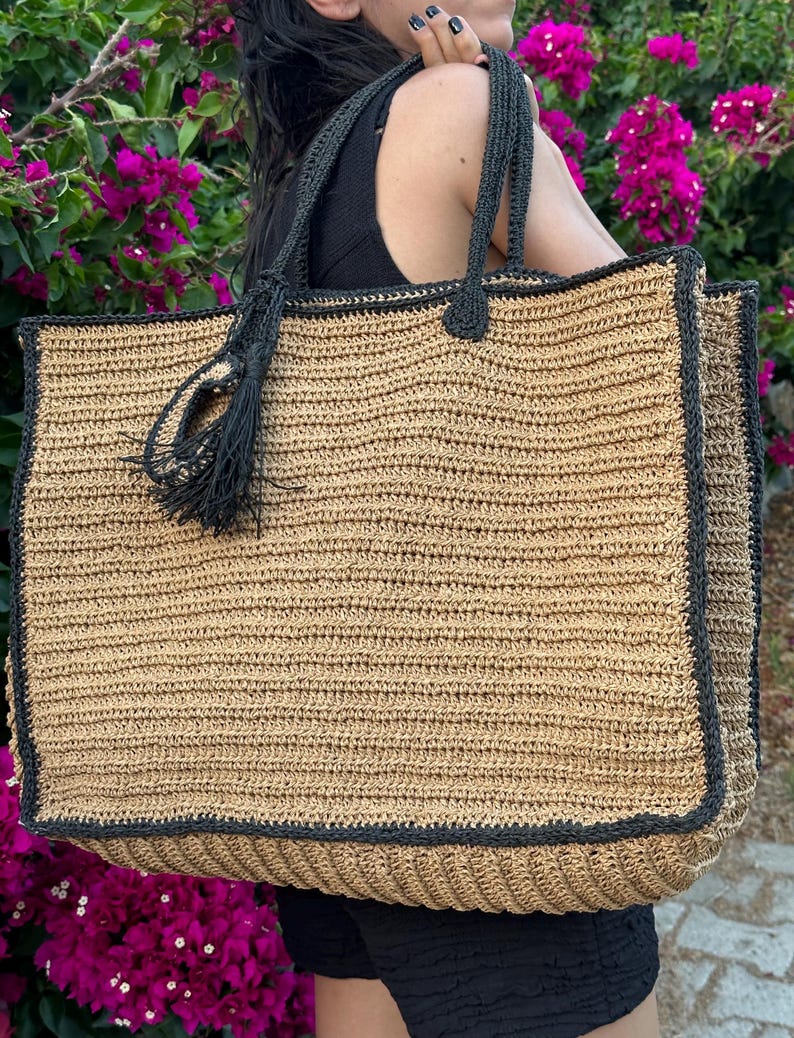 K&ouml;nnte beinhalten: Eine gro&szlig;e, gewebte Tragetasche mit schwarzem Besatz und einem schwarzen Quasten. Die Tasche ist aus einem nat&uuml;rlichen stroh&auml;hnlichen Material gefertigt.