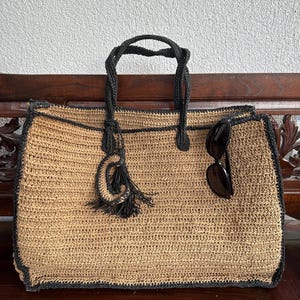 K&ouml;nnte beinhalten: Eine gro&szlig;e, beige geh&auml;kelte Tragetasche mit schwarzem Besatz und einem schwarzen Quasten. Die Tasche steht auf einer Holzoberfl&auml;che und enth&auml;lt eine Sonnenbrille.