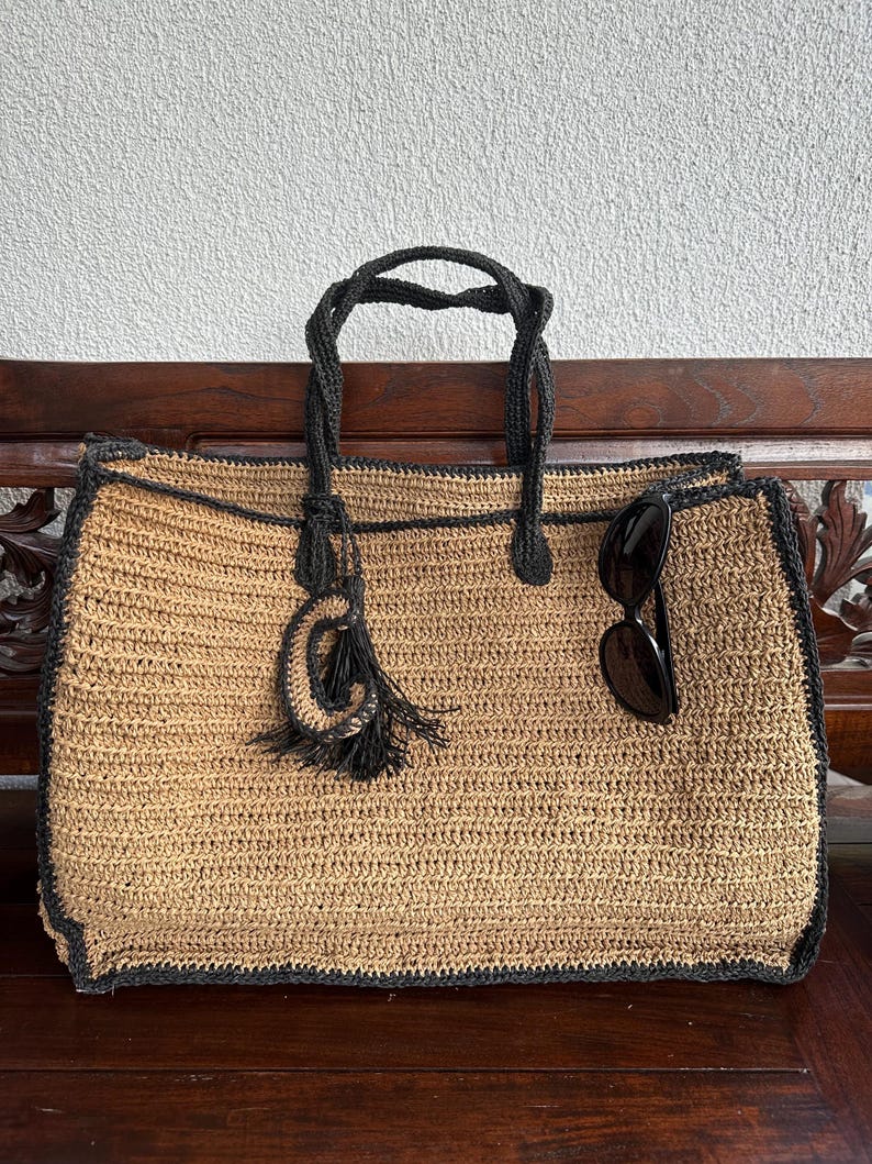 K&ouml;nnte beinhalten: Eine gro&szlig;e, beige geh&auml;kelte Tragetasche mit schwarzem Besatz und einem schwarzen Quasten. Die Tasche steht auf einer Holzoberfl&auml;che und enth&auml;lt eine Sonnenbrille.