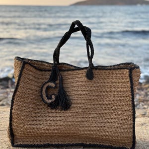 K&ouml;nnte beinhalten: Eine gro&szlig;e, braune geh&auml;kelte Tragetasche mit schwarzem Besatz und einem schwarzen Quasten. Die Tasche hat einen gro&szlig;en, schwarzen Buchstaben "G" auf der Vorderseite.