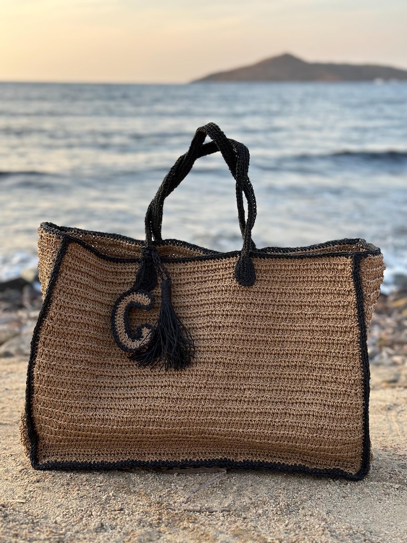 K&ouml;nnte beinhalten: Eine gro&szlig;e, braune geh&auml;kelte Tragetasche mit schwarzem Besatz und einem schwarzen Quasten. Die Tasche hat einen gro&szlig;en, schwarzen Buchstaben "G" auf der Vorderseite.