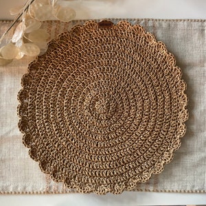 Pode incluir: Um sousplat redondo e tecido, com uma borda ondulada, feito de fibras de cor natural. O sousplat é colocado em cima de um sousplat retangular de linho bege claro com uma borda costurada. Uma pequena etiqueta marrom está anexada.