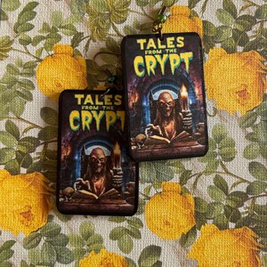 May include: A pair of rectangular dangle earrings featuring the Tales from the Crypt logo in yellow and green text above an illustration of a skeletal figure holding a candle while reading a book, attached to gold-toned metal earring hooks.