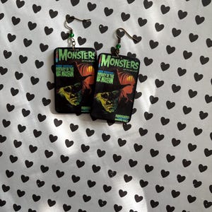 May include: A pair of earrings featuring a vintage movie poster design. The earrings display the text "MONSTERS" and "Mystery of the Wax Museum" with a green and orange color scheme. The earrings have a hook closure and green beads.