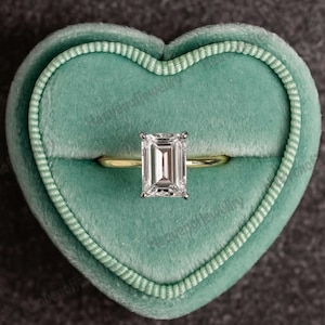Puede incluir: Un anillo de diamantes con talla esmeralda y banda de oro descansa en un joyero de terciopelo verde claro en forma de corazón. El joyero está sobre una superficie gris oscuro. El texto "2.75ct" está en la parte inferior de la imagen.