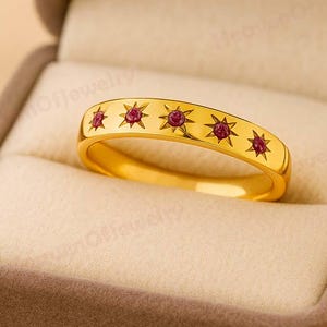 Pode incluir: Um anel de ouro com um design de estrela e rubi, apresentado numa caixa de joias castanha. O anel apresenta uma série de estrelas douradas, cada uma com um pequeno rubi redondo no centro. O anel é de cor dourada brilhante.
