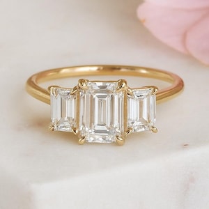 May include: A gold ring with three emerald-cut diamonds. The center diamond is larger than the two side stones. The ring is set on a white surface, with a pink flower in the background.