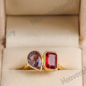 Könnte beinhalten: Ein goldener Ring mit zwei Edelsteinen: einem birnenförmigen lila Stein und einem rechteckigen roten Stein. Der Ring ist in einem goldenen Band gefasst und in einer beigen Samtringbox präsentiert.