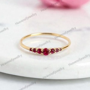 Könnte beinhalten: Ein zarter Goldring mit einer Reihe leuchtend roter Edelsteine. Das schlanke Band ergänzt die satte Farbe der Steine und schafft ein klassisches und elegantes Design. Der Ring liegt auf einer weißen Marmoroberfläche.