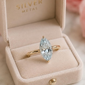 Peut inclure: Une bague en or avec une pierre précieuse bleu clair de taille marquise, présentée dans un écrin en velours beige ouvert. La boîte porte l'inscription "SILVER METAL" en lettres dorées. Des roses roses sont en arrière-plan.