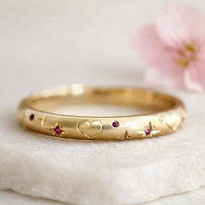 Può includere: Un anello d'oro con cuori, stelle e punti incisi, impreziosito da piccole gemme rosse. L'anello è presentato su una superficie bianca, con un fiore rosa tenue sullo sfondo. Il design dell'anello è delicato e romantico.