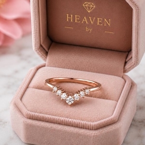 May include: A rose gold V-shaped ring featuring seven small round clear stones set along the band. The ring is displayed inside an open, dusty pink velvet jewellery box with the text HEAVEN by printed in gold on the inner lid of the box.