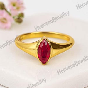 Puede incluir: Un anillo de oro con una piedra preciosa roja oscura con corte marquesa. El anillo tiene una banda lisa y pulida y un engaste que sujeta la piedra preciosa de forma segura. El anillo se exhibe sobre una superficie blanca.