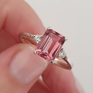 May include: A silver ring with a pink gemstone and two smaller diamonds on the band.