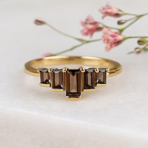 May include: A gold ring featuring five rectangular, smoky brown gemstones. The gemstones are set in a row, with the center stone slightly larger. The ring is set against a white marble surface with pink flowers in the background.