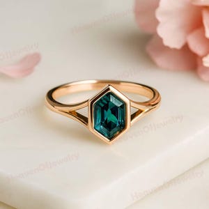 Peut inclure: Une bague en or avec une pierre précieuse hexagonale de couleur teal. Le bracelet de la bague se divise en deux fines griffes de chaque côté de la pierre précieuse, créant un design ouvert. La bague est posée sur un fond blanc doux.