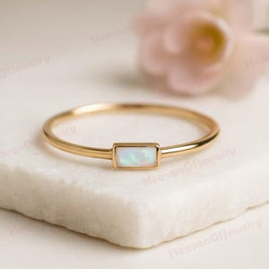 May include: A delicate gold ring featuring a rectangular opal stone. The ring has a thin band and a minimalist design. The opal stone displays a play of colors, set in a simple bezel setting. The ring is displayed on a white marble surface.