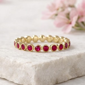 May include: A gold-colored ring featuring a series of vibrant red gemstones. The gemstones are evenly spaced around the band, creating a continuous pattern. The ring is set on a white marble surface, with soft pink flowers in the background.