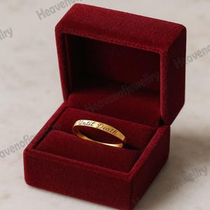 May include: A gold band ring with the inscription "Until Death" inside a red velvet ring box. The ring is centered in the open box, which is a deep red color. The ring's inscription is in a cursive font.