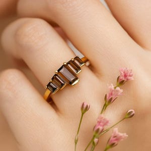 May include: A gold ring featuring five rectangular, brown gemstones. The ring is set on a gold band and is displayed on a person's finger. Pink flowers are in the background.