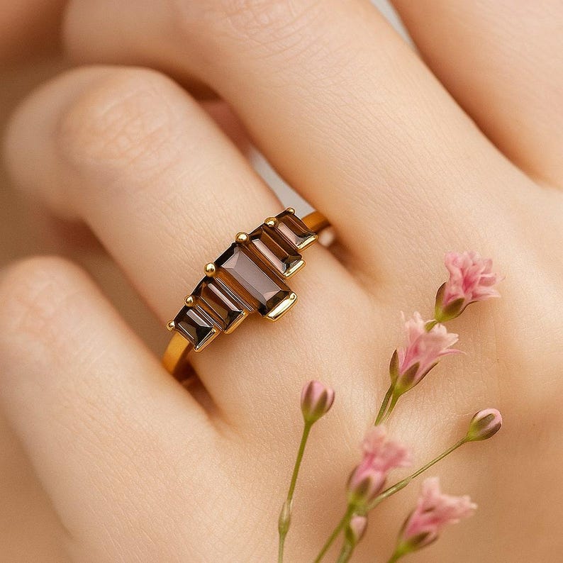 May include: A gold ring featuring five rectangular, brown gemstones. The ring is set on a gold band and is displayed on a person's finger. Pink flowers are in the background.