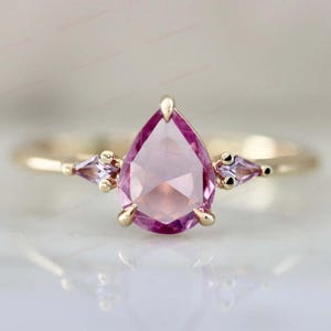May include: A gold ring featuring a large, pear-shaped pink gemstone as the centerpiece, flanked by two smaller, triangular purple gemstones. The ring has a delicate band and a polished finish, showcasing a classic design.