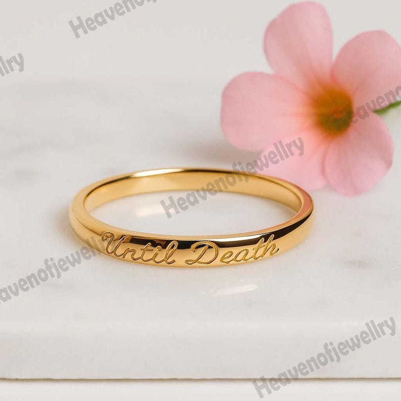 May include: A gold-colored ring with the inscription "Until Death" engraved on the band. The ring is set against a white marble surface, with a pink flower in the background. The ring is a simple band style.