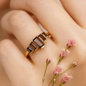 May include: A gold ring featuring five rectangular, brown gemstones. The ring is set on a gold band and is displayed on a person's finger. Pink flowers are in the background.