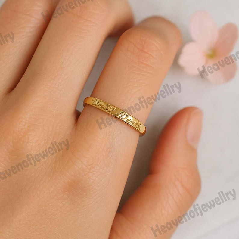 May include: A gold-colored ring with the inscription "Until Death" engraved on the outside. The ring is on a finger, with a soft-focus background.