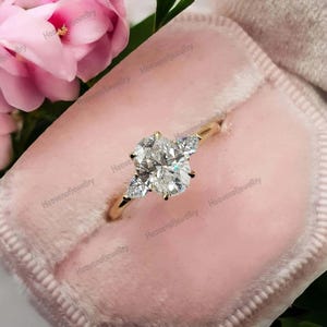 May include: A gold ring featuring a large oval-cut diamond and two pear-shaped side stones. The ring is set in a delicate band and presented in a pink velvet ring box. Pink flowers are visible in the background.