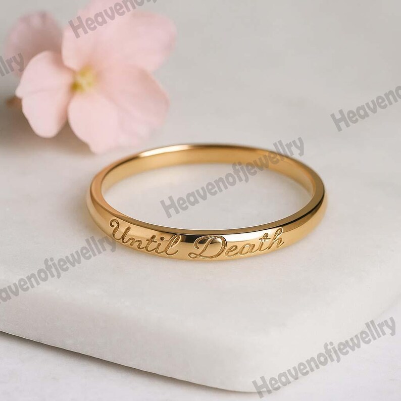 May include: A gold-colored ring with the inscription "Until Death" engraved on the band. The ring is set against a white surface, with a soft pink flower in the background. The ring is a simple band design.