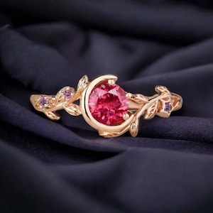 May include: A gold-colored ring with a large, teardrop-shaped, red gemstone as the focal point. The ring features a crescent moon design around the gemstone and is adorned with leaf and small purple gemstone accents. The ring is set against a dark blue background.