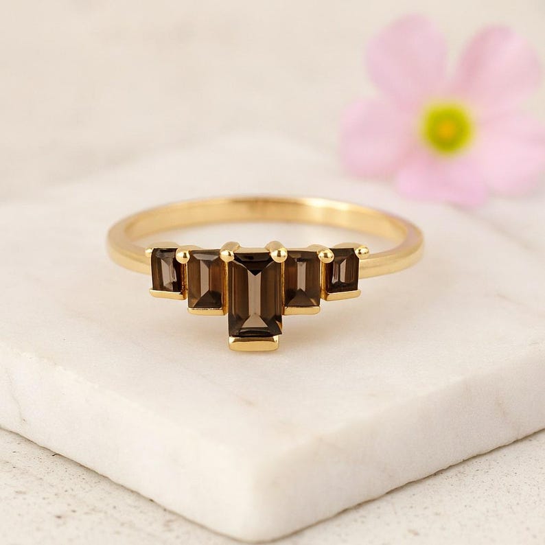 May include: A gold ring featuring five rectangular, dark brown gemstones. The gemstones are arranged in a tiered pattern, with the largest stone in the center. The ring is set on a white surface, with a blurred pink flower in the background.