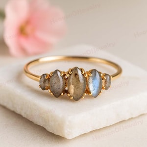 May include: A gold ring with five oval gemstones. The gemstones display grey, brown, and blue tones. The ring is set on a white surface. A soft pink flower is visible in the background.