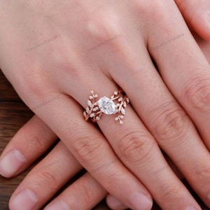 Puede incluir: Juego de anillos de compromiso de oro rosa con una piedra central de diamante de talla ovalada. La banda está adornada con delicados detalles de hojas y vides, y pequeñas piedras de acento. Una banda a juego completa el conjunto.