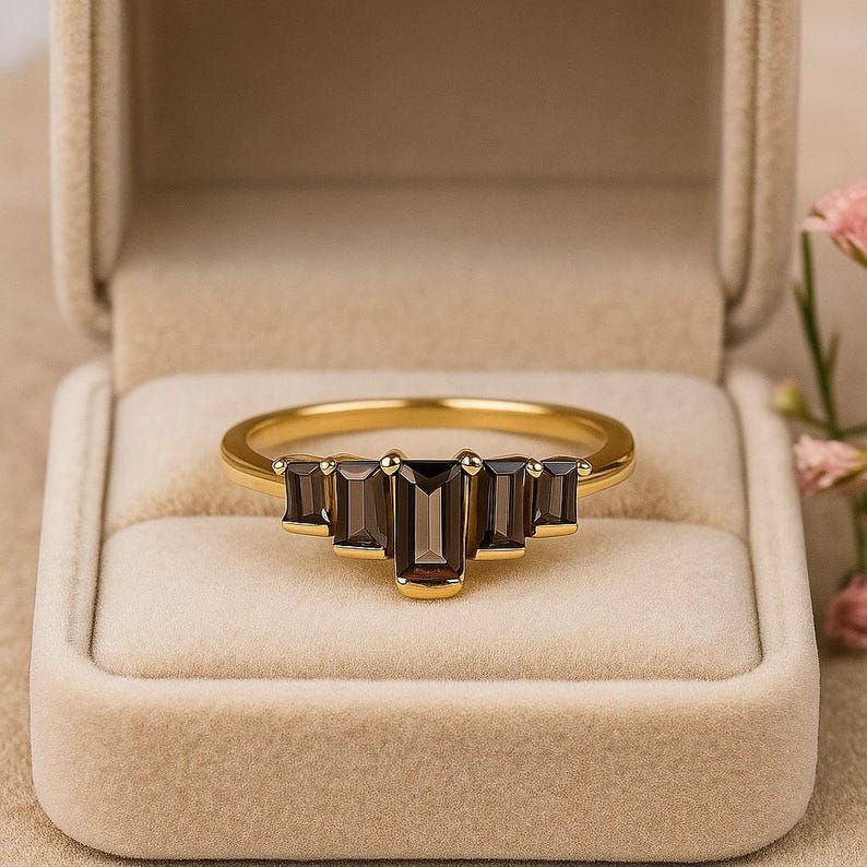 May include: A gold ring with five rectangular, dark brown gemstones set in a row. The ring is displayed in a beige jewelry box, with a soft focus background.