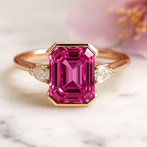 Może przedstawiać: Pierścionek z różowego złota z dużym, prostokątnym, fuksjowym kamieniem szlachetnym w złotej oprawie. Dwa przezroczyste kamienie szlachetne w kształcie gruszki flankują centralny kamień. Obręcz pierścionka jest z polerowanego złota.