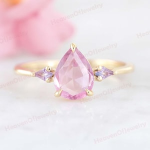 以下が含まれることがあります： 中央に洋梨型のピンク色の宝石をあしらったゴールドリング。両側には、2つの小さな三角形の紫色の宝石が配置されています。リングのバンドはシンプルな磨かれたゴールドで、宝石は繊細な爪でセットされています。